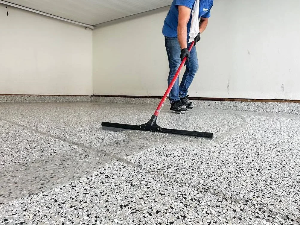 Worker applying gray speckled epoxy coating to concrete floor with squeegee tool